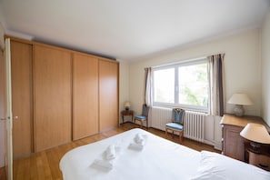 A hotel room with a large bed, wooden wardrobe, two chairs, a table with a lamp, and a window with curtains. - Maison Bellevue, private terrace with view (Brunstatt-Didenheim)