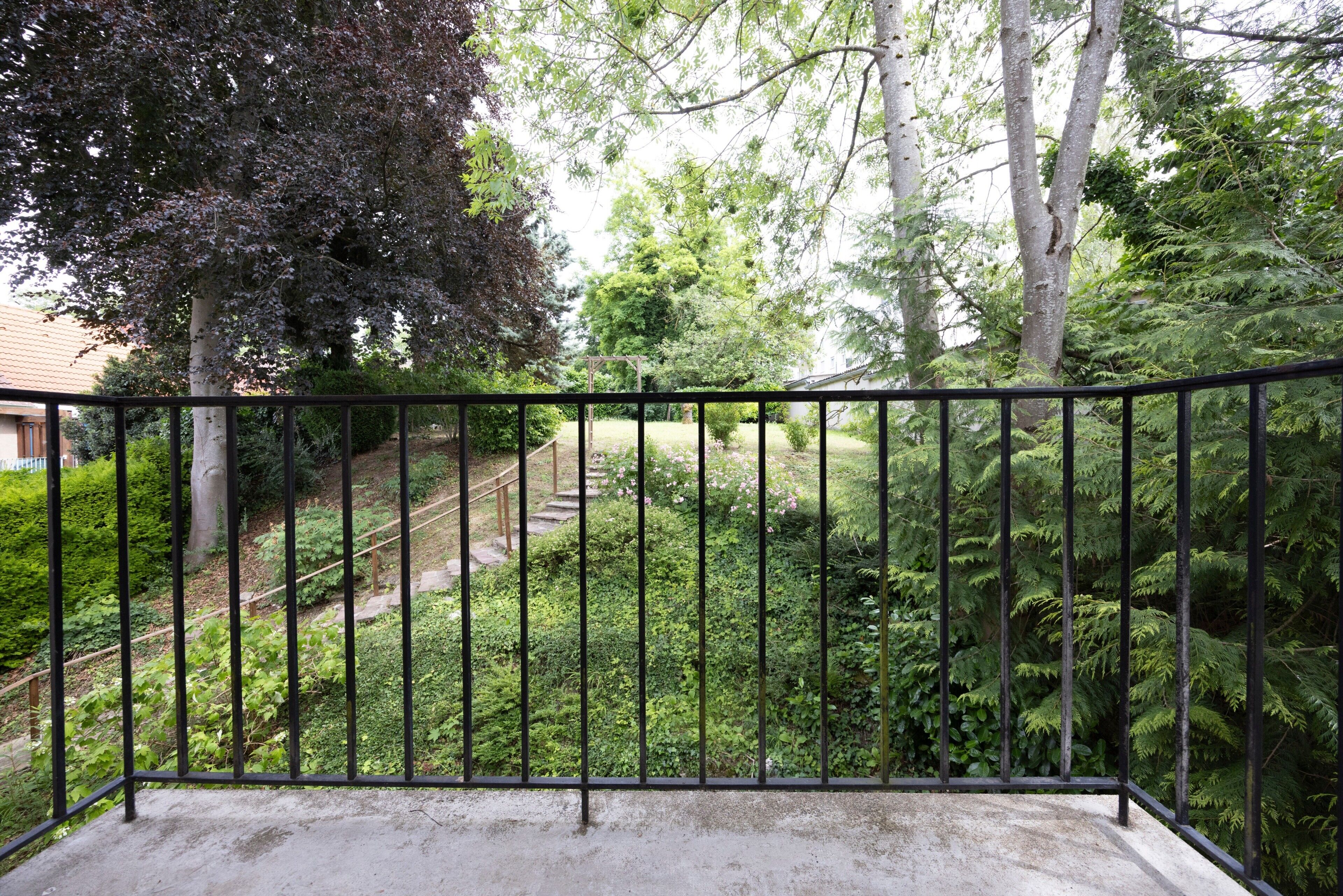 A balcony with a metal railing overlooking a garden with trees and a staircase.