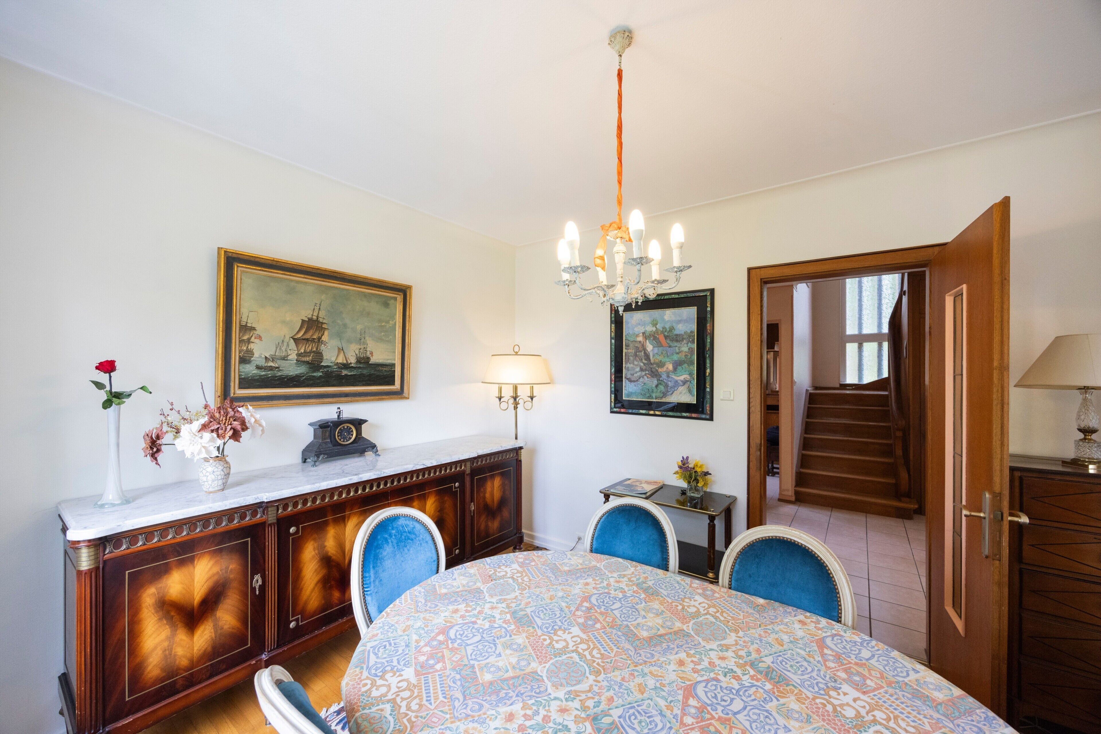 A dining area with a patterned tablecloth, a chandelier, a cabinet with a vase of flowers, and a painting on the wall.