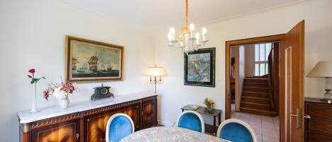 A dining area with a patterned tablecloth, a chandelier, a cabinet with a vase of flowers, and a painting on the wall.