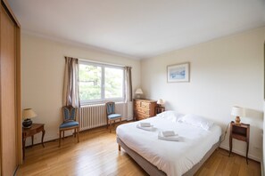 Ein Schlafzimmer mit Bett, zwei Stühlen, einem Schminktisch, einem Nachttisch, einem Fenster mit Vorhängen und einer Heizung.