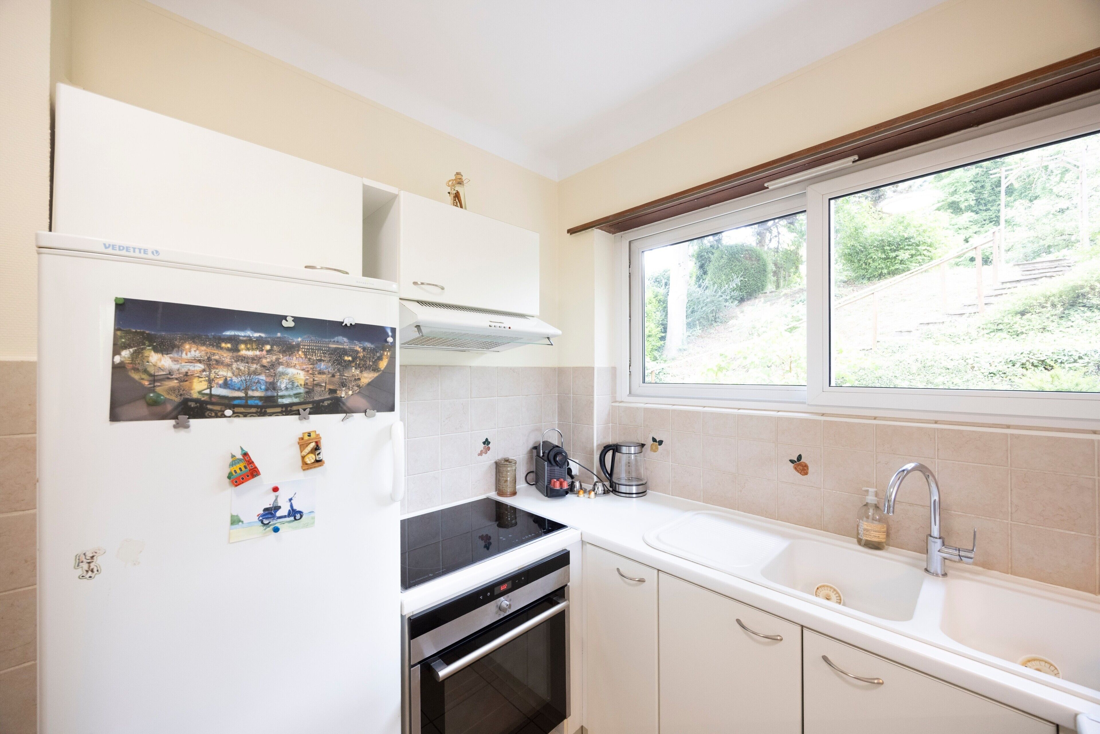 A kitchen with a built-in oven, a stovetop, a sink, and a window with a view of greenery.
