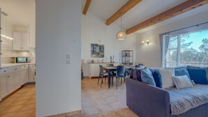 Una cocina moderna con armarios blancos, un comedor con mesa y sillas, un sofá con cojines y un amplio ventanal con vistas a árboles.