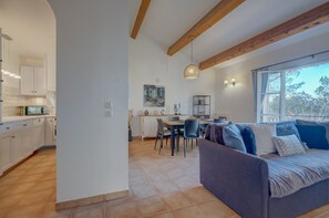 Eine moderne Küche mit weißen Schränken, ein Essbereich mit Tisch und Stühlen, eine Sitzecke mit Kissen sowie ein großes Fenster mit Blick auf Bäume.