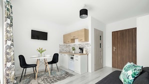 A modern kitchen with white cabinets, a black countertop, and a built-in oven. There is a dining area with a table and chairs, and a large window with a view of the city. - Cozy Studio close to the Beach | Balcony | Parking (Grzybowo)