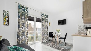 A modern living room with a sofa, a dining area with a table and chairs, a kitchen area with a countertop and cabinets, and a sliding glass door leading to a balcony. - Cozy Studio close to the Beach | Balcony | Parking (Grzybowo)