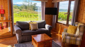 A cozy room with a sofa, armchair, and a TV. There's a window with a view of greenery and a fire extinguisher on the wall.