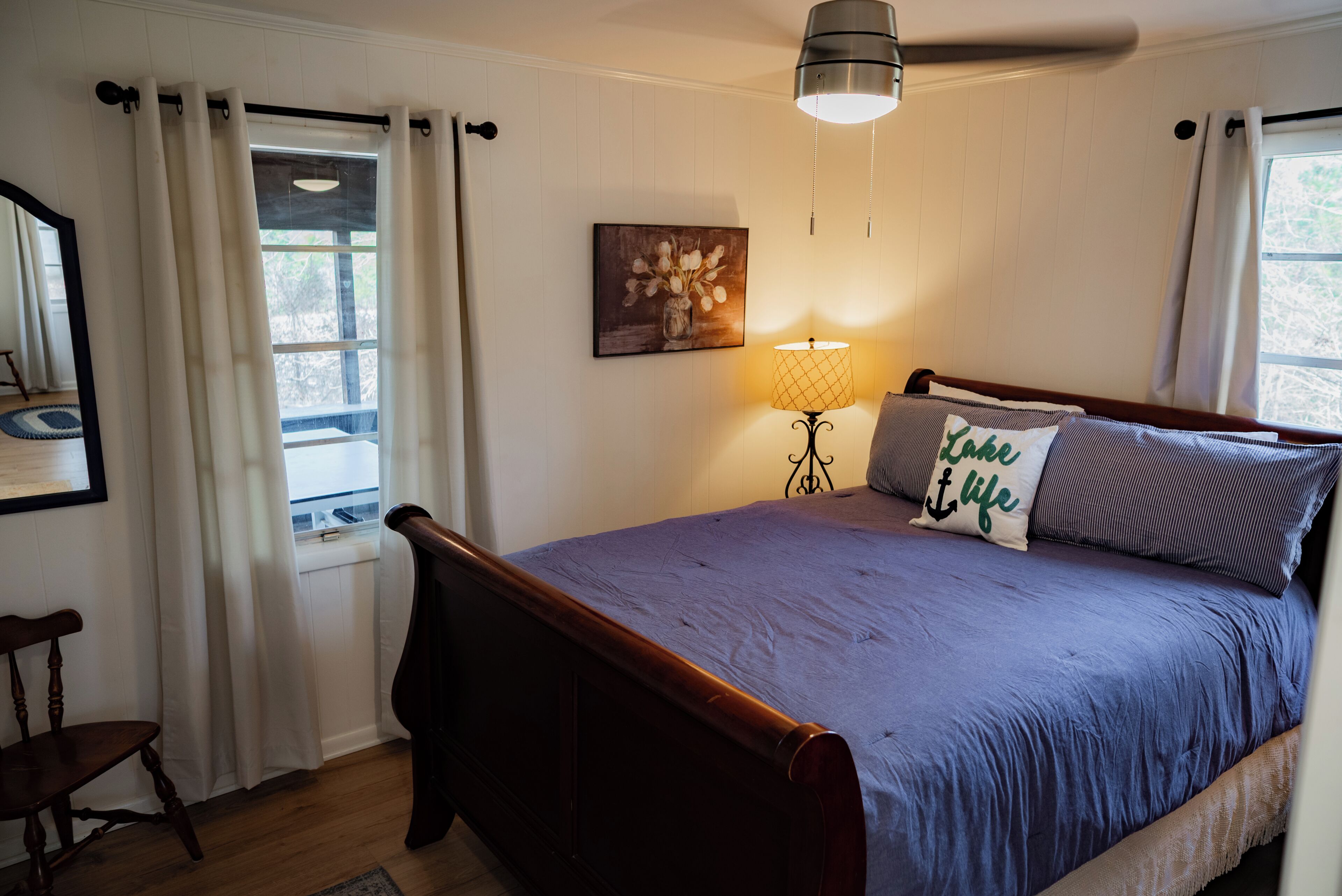 Ein Schlafzimmer mit einem hölzernen Bett, einem fensterbelaubten Fenster, einer Wandlampe und einem gerahmten Bild.