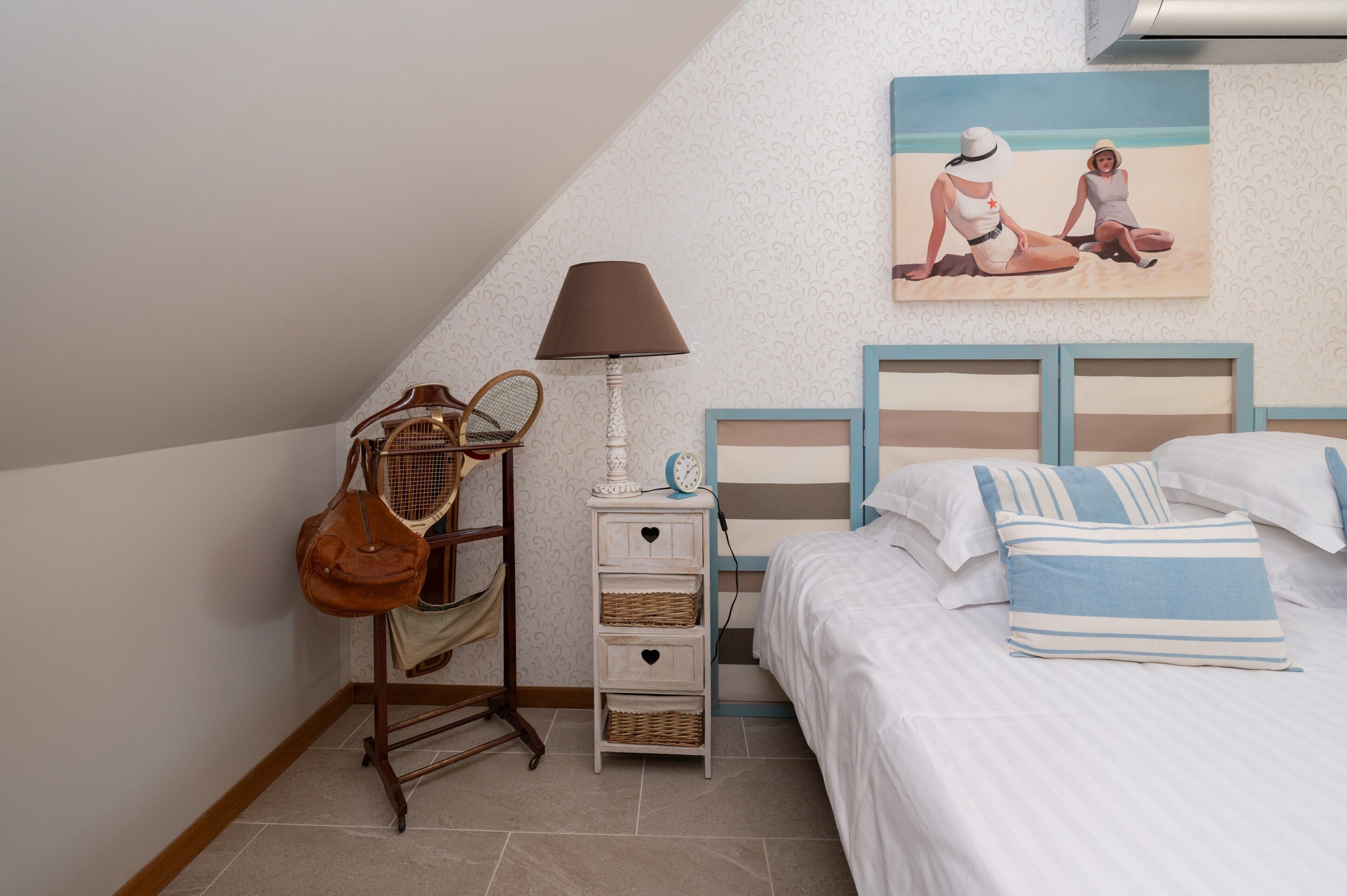 Ein Schlafzimmer mit Bett, Nachttisch, Lampe, einem Korbwägelchen und einem Gemälde von zwei Personen am Strand.