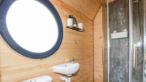 Bany d'una cabana de fusta amb una finestra rodona, un lavabo, un vàter i una dutxera.