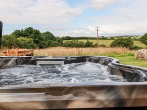 Un jacuzzi con banco de madera y una mesa, rodeado por un campo de césped y árboles.
