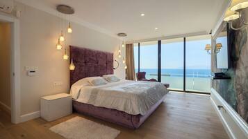 A bedroom with a large bed, bedside tables, a fluffy rug, and a view of the sea through large windows.