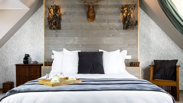 Un dormitorio con cabecera de madera, cama con ropa de cama blanca y oscura, mesita de noche con lámpara y una silla con cojín.