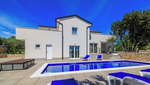 Una casa moderna con piscina, sillones de descanso azules y un muro de piedra.