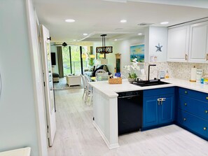 A modern kitchen with blue cabinets, a dishwasher, and a dining area with a view. - Island Club 106 | Pool, Hot Tub, Tennis & Beach (Hilton Head Island)