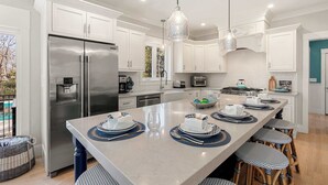 A modern kitchen with white cabinets, stainless steel appliances, a central island with seating, pendant lighting, and a view of the outdoors. - Upscale Beach Retreat w Pool, Half Mile to Beach! (Dennis)