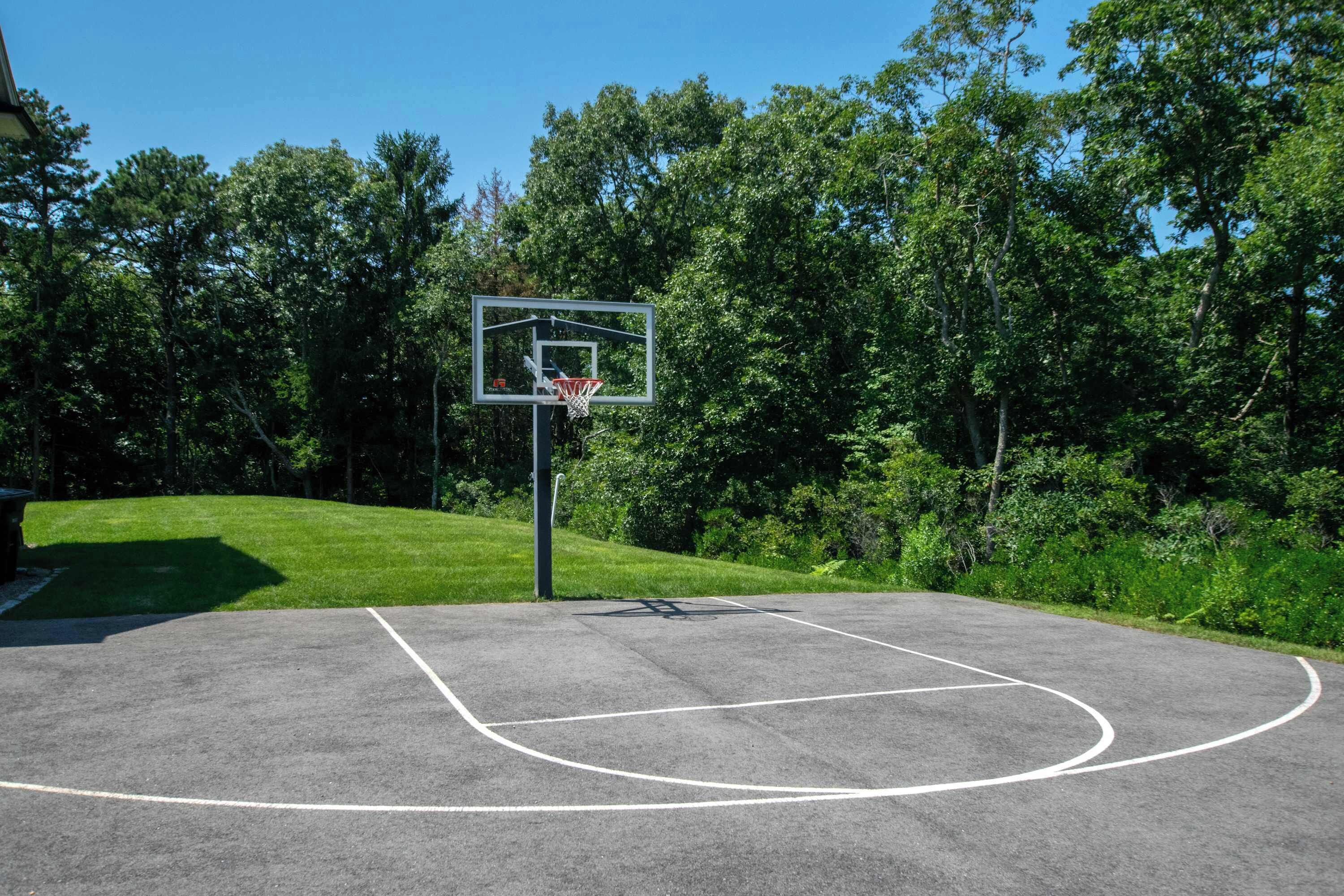 An outdoor basketball court with a hoop, surrounded by trees and grass.