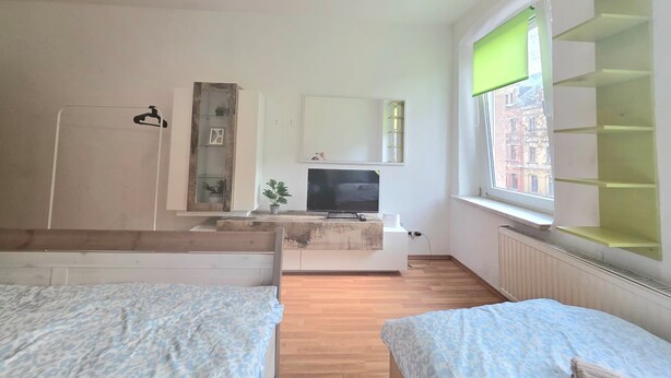 A modern bedroom with a bed, a TV, a shelf with plants, and a window with a view of buildings. - Cozy 1 bedroom apartment in a quaint Chemnitz neighborhood (Chemnitz)