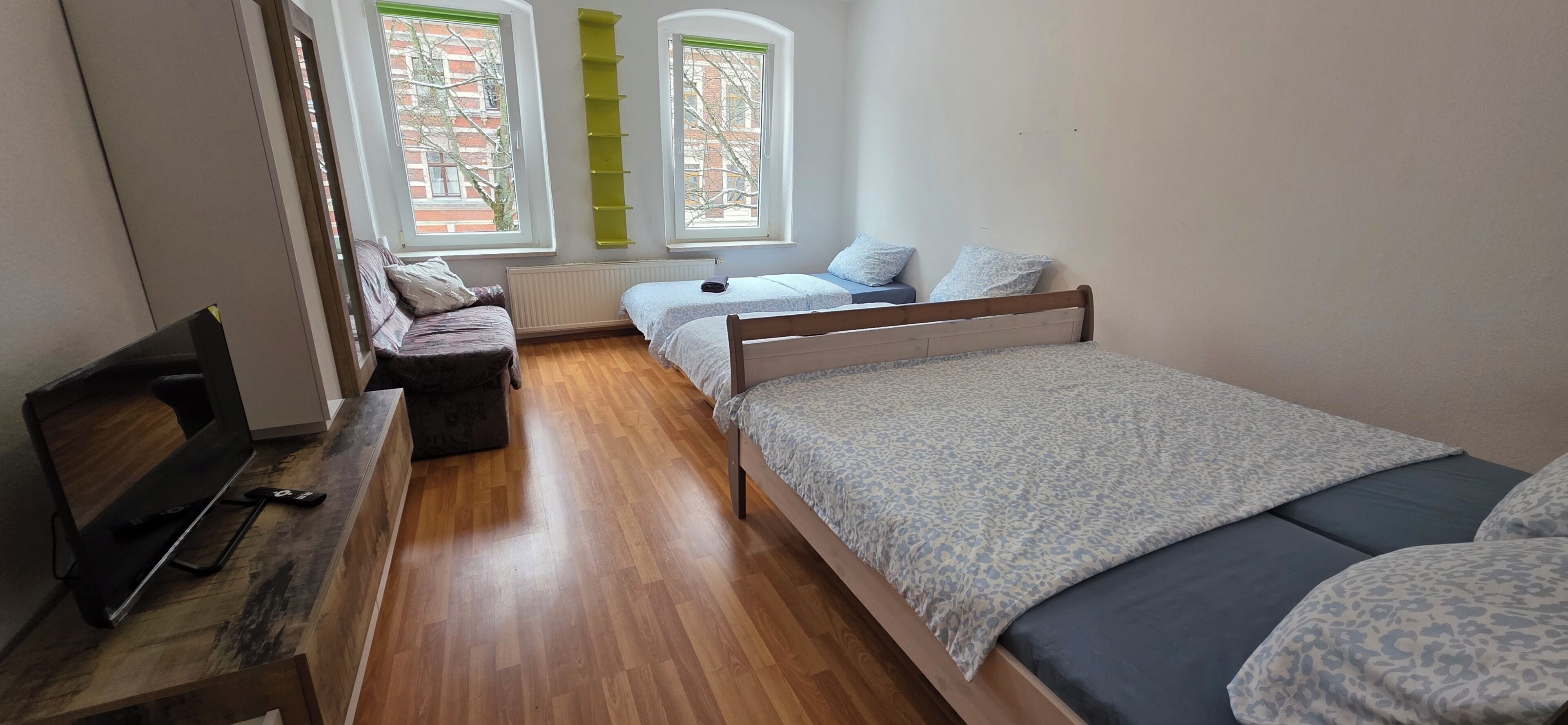 A bedroom with two beds, a wooden desk, a TV, and a window with a view of buildings.
