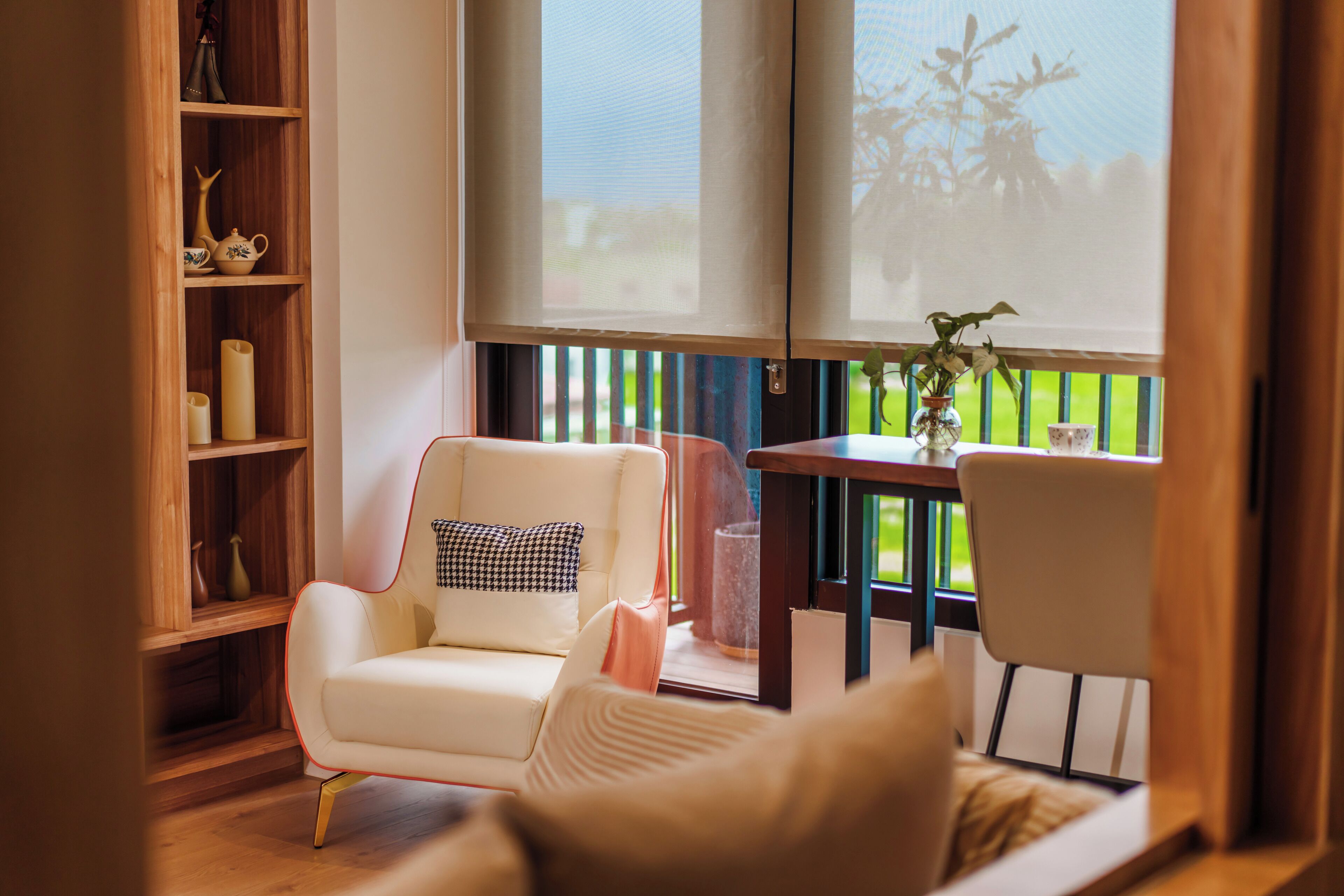 A room with a white sofa, a wooden bookshelf, a small table with a vase, and a view through a window.
