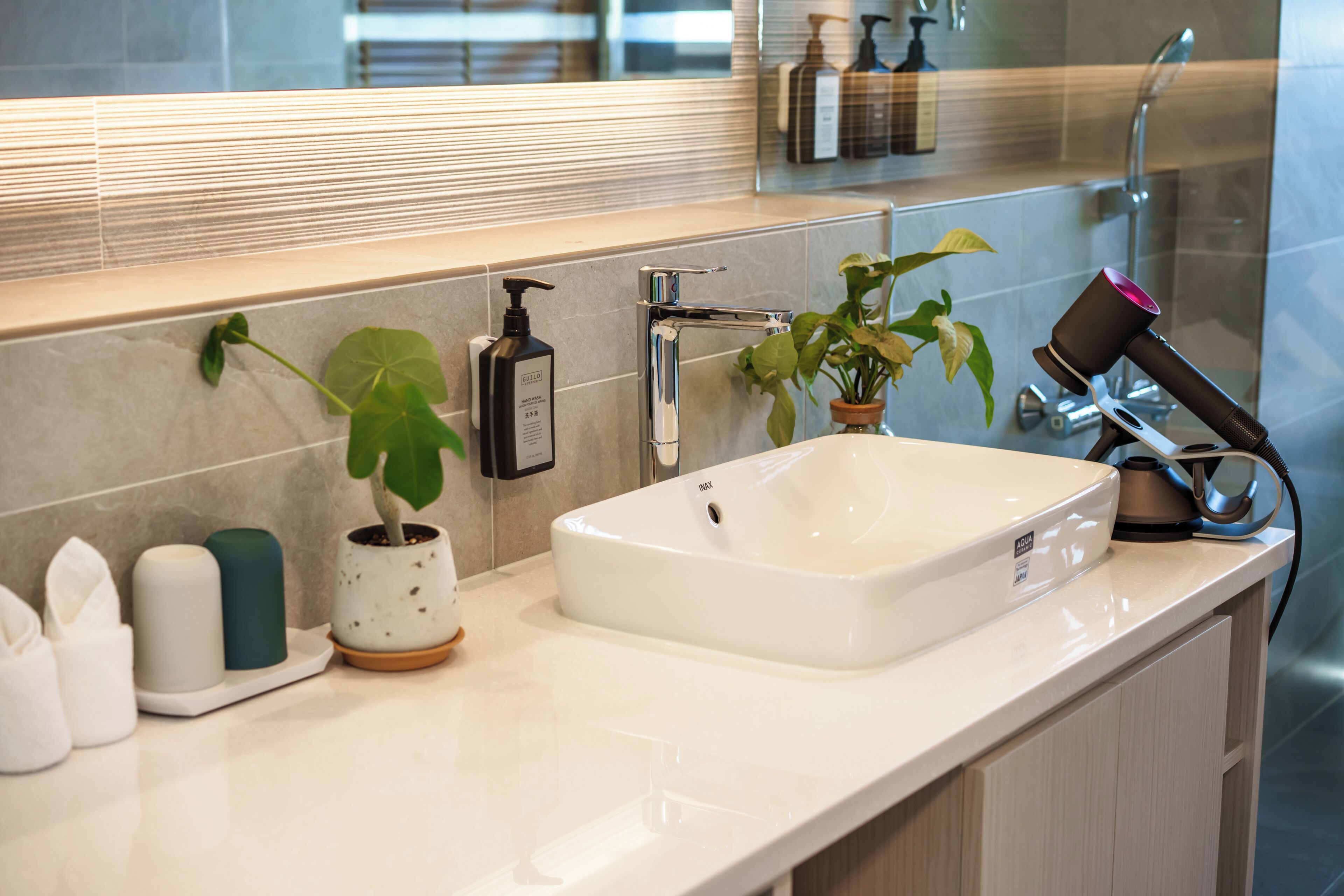 Une salle de bain avec un lavabo blanc, un miroir, une plante en pot, un sèche-cheveux et des articles de toilette posés sur le comptoir.