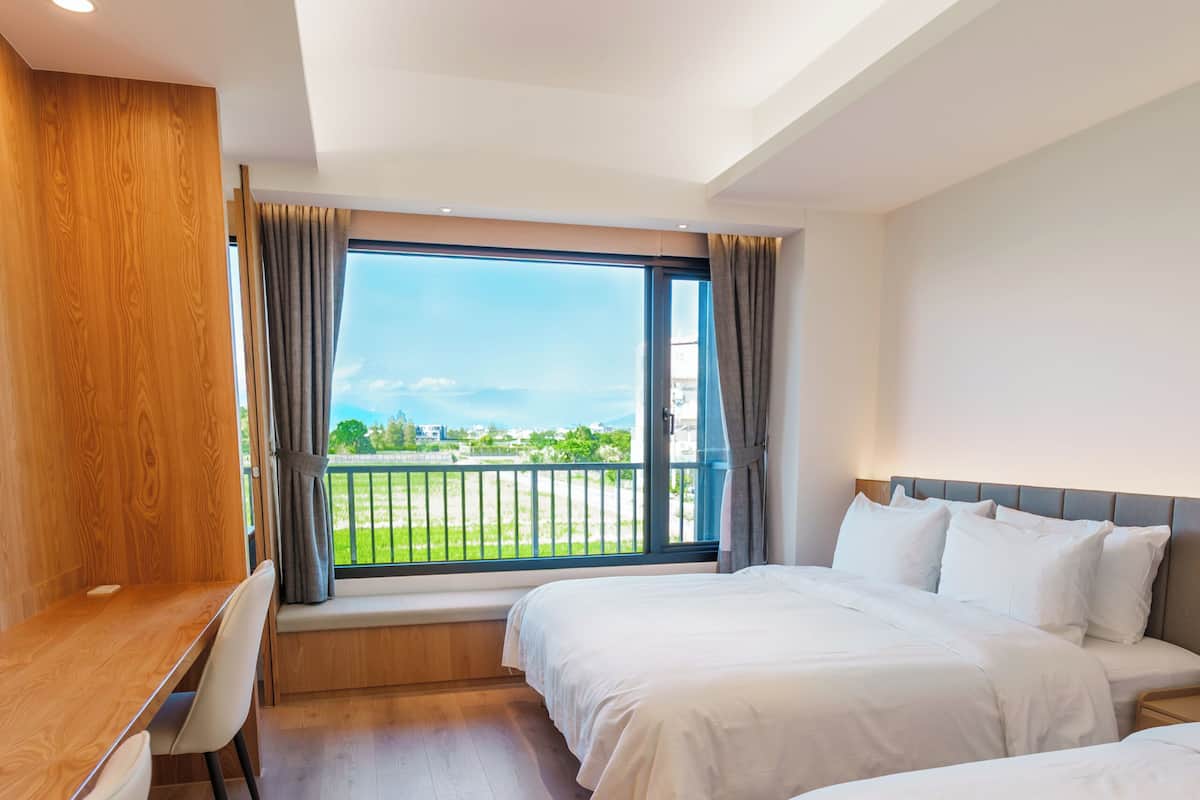 A hotel room with a wooden desk, two beds, a window with a view, and a balcony.