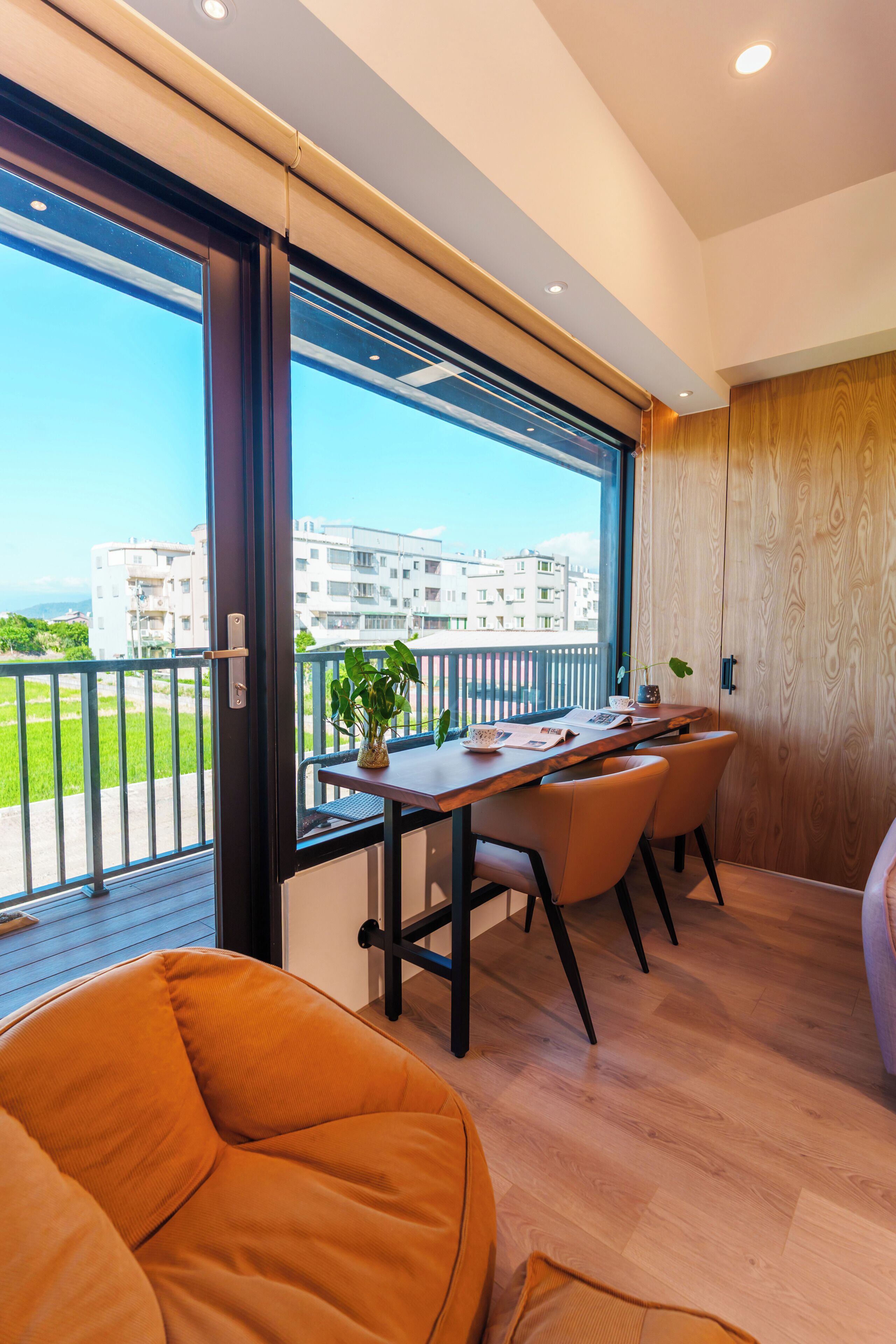 A modern room with a wooden floor, a dining table with chairs, a sofa, and a large window with a view of buildings and a green area.