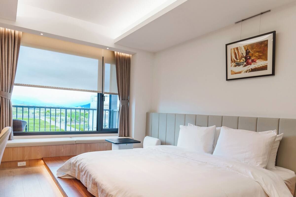 A bedroom with a large bed, a window with curtains, a painting on the wall, and a view of the outdoors.