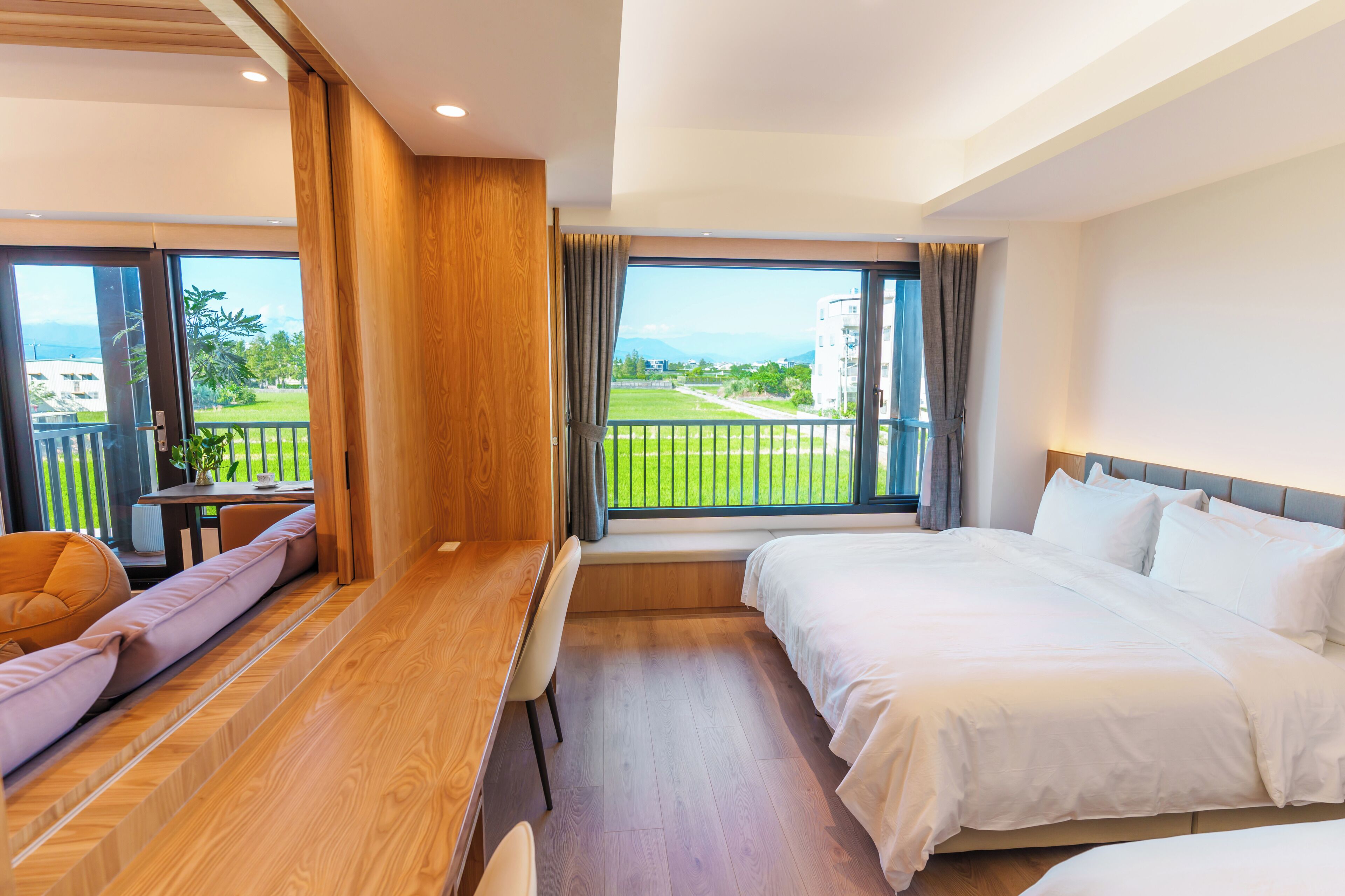 A hotel room with a bed, a desk, a chair, a window with a view of a green landscape, and a balcony.