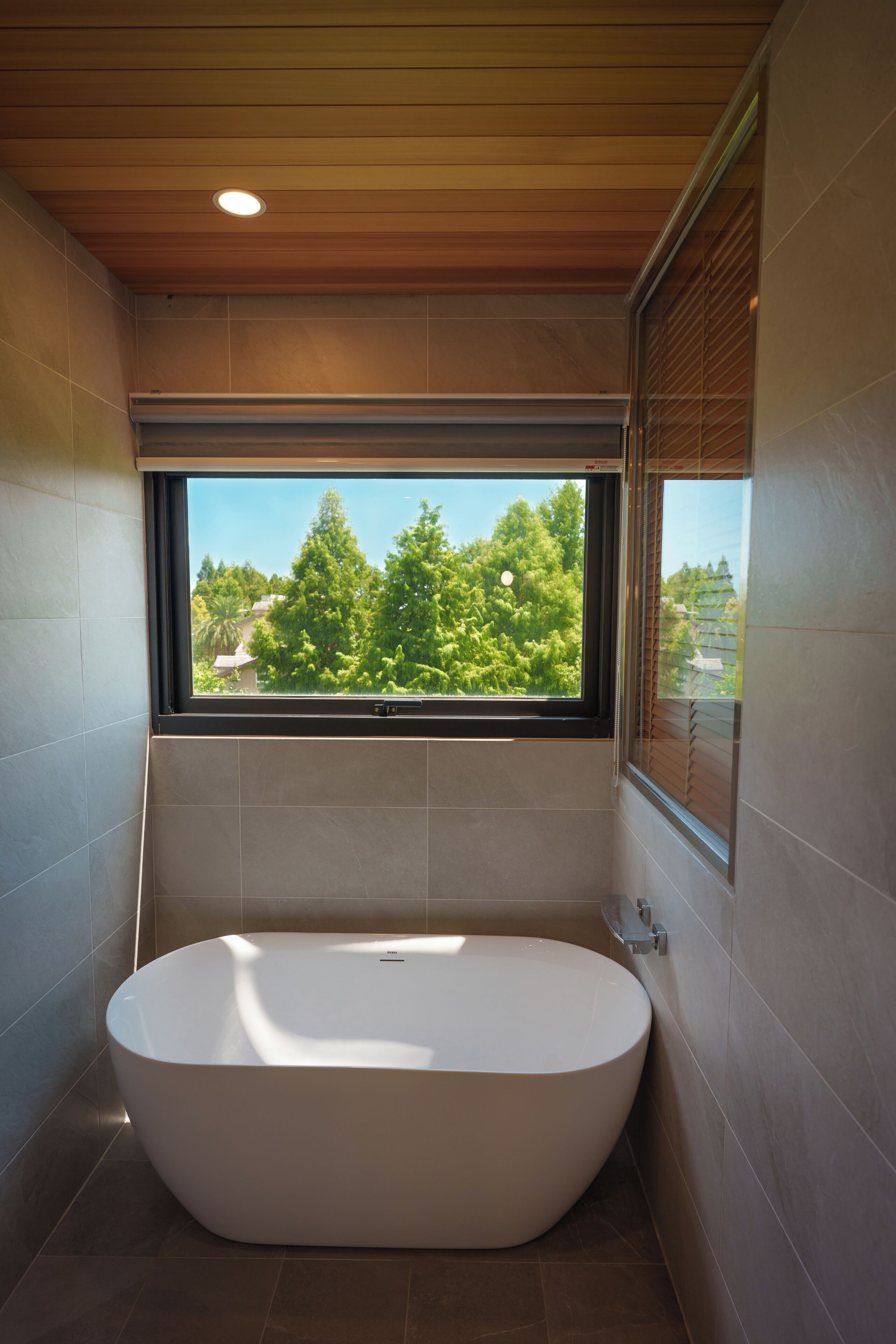 Une salle de bain moderne avec une baignoire sur pieds, une fenêtre donnant sur la verdure et des panneaux de plafond en bois.