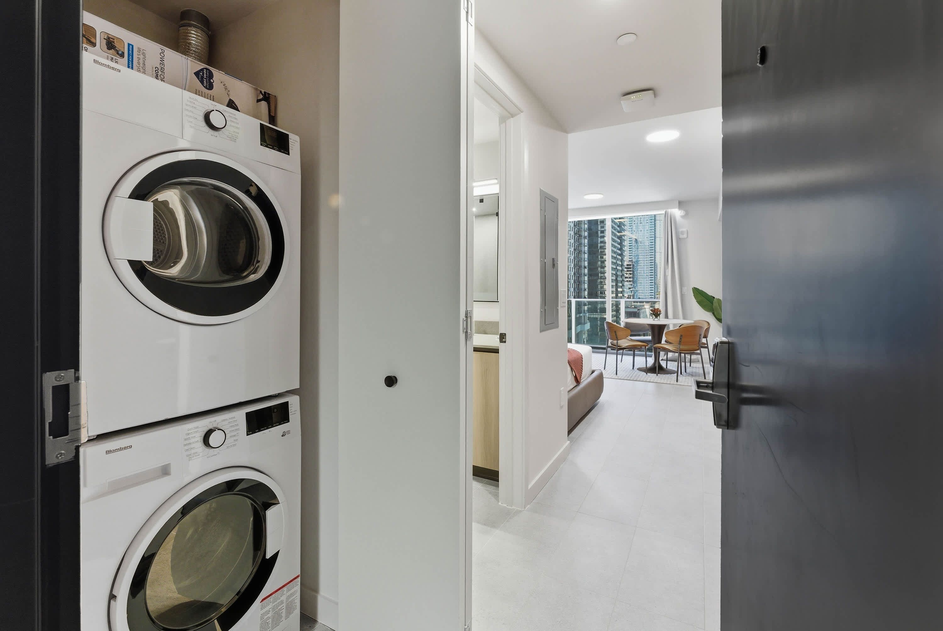 A laundry room with stacked washing machines, a dining area with chairs, and a view of high-rise buildings.