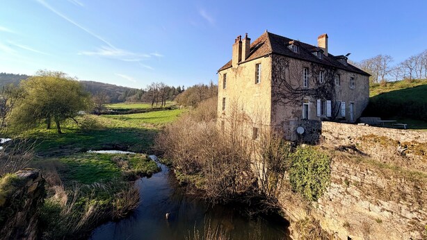 A stone building with a chimney, a small stream, and a grassy field. - Former mill of the Château de Bourbilly transformed into a pretty country house (Vic-de-Chassenay)