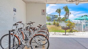 Une station de location de vélos avec des vélos à disposition, un panneau indiquant l’interdiction de se garer, et une vue sur un bâtiment bordé de palmiers et entouré d’une clôture blanche.