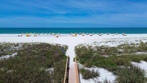 A beach with a wooden pathway leading to the shore, surrounded by sand dunes and beach umbrellas. - Sunset Vistas 302n (Treasure Island)
