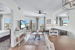 A modern living room with a dining area, large windows, ceiling fan, and a flat-screen TV. - Sunset Vistas 605n (Treasure Island)