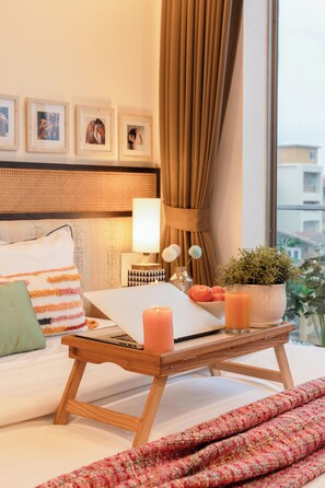 Habitación de hotel ordenada con una cama, una mesita de noche con un ordenador portátil, una lámpara, una planta en maceta y una ventana con cortinas.