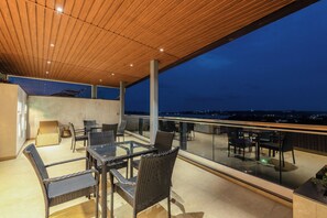 A rooftop terrace with glass balustrades, wicker furniture, and a view of the city at dusk. - Staymaster Flabris 3BHK Apartment (Panaji)