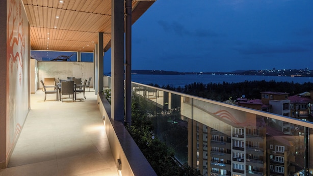 Un balcón con vistas al mar y edificios, techo de madera y zona de comedor con sillas y mesas.