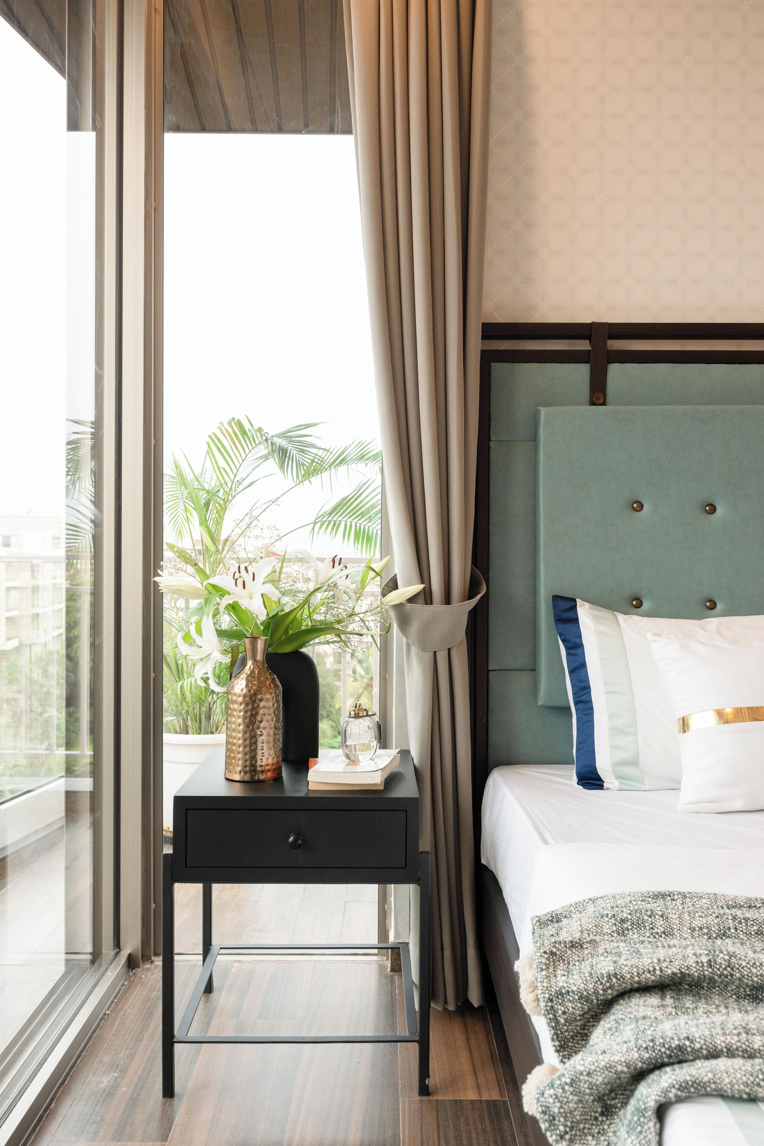 A hotel room with a bed, a bedside table with a vase of flowers, a window with curtains, and a view of buildings outside.