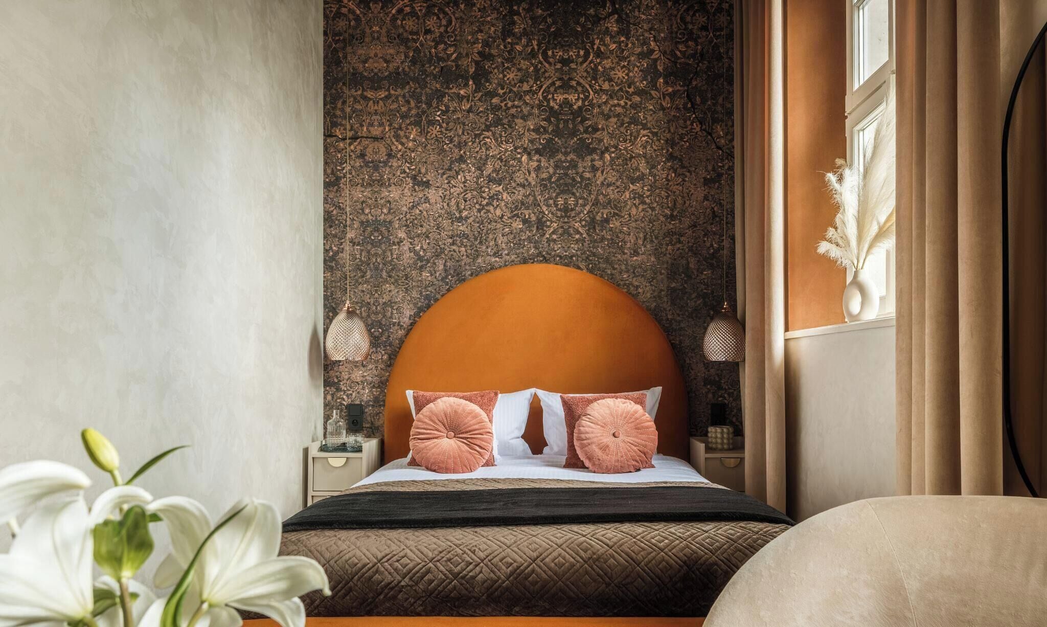 A bedroom with a large bed, a textured wall, a nightstand, a vase with dried flowers, and a window with curtains.