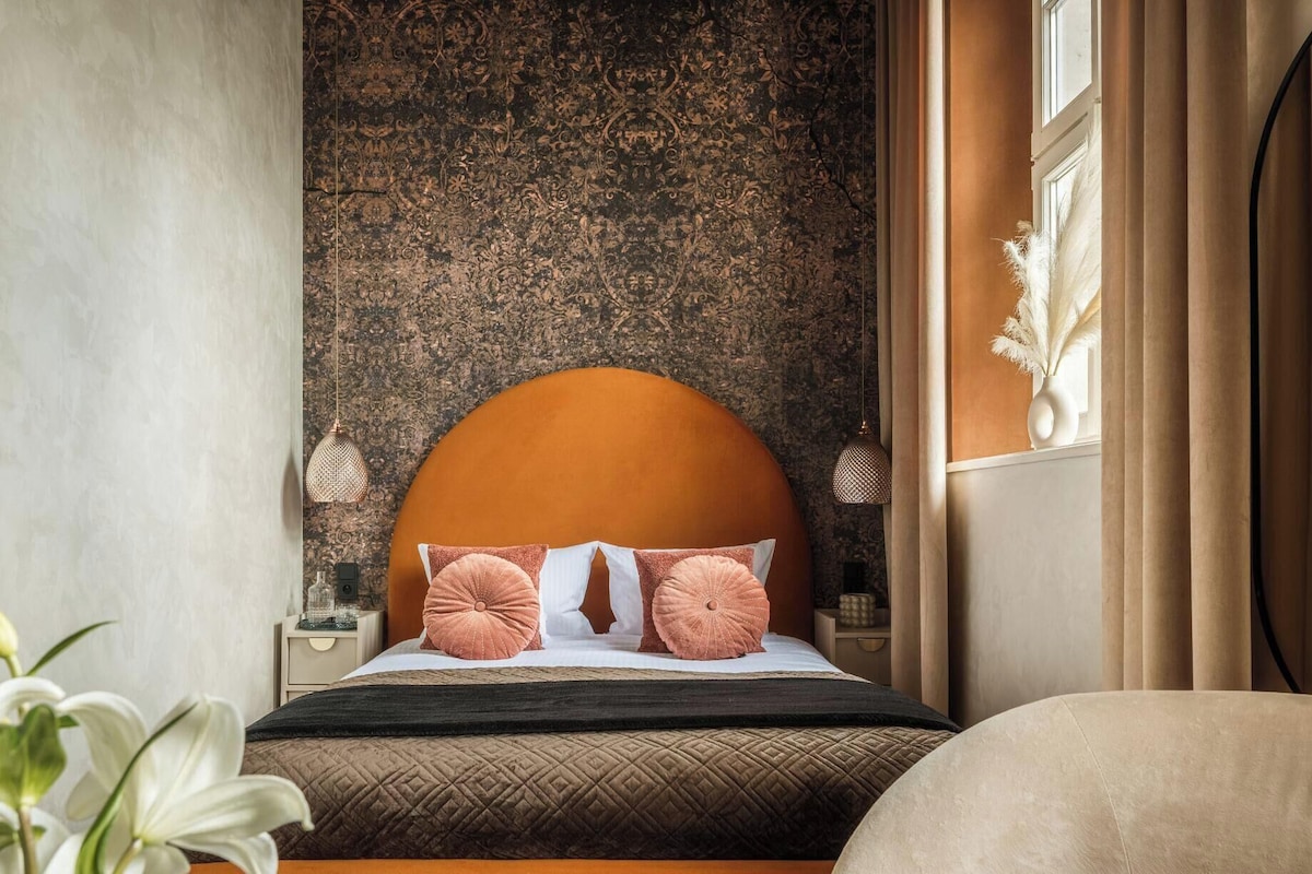 Un dormitorio con una cama grande, una pared con textura, una mesita de noche, un jarrón con flores secas y una ventana con cortinas.