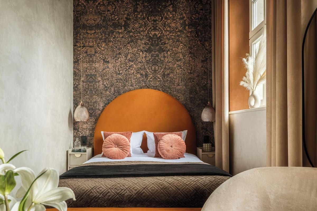 A bedroom with a large bed, a textured wall, a nightstand, a vase with dried flowers, and a window with curtains.