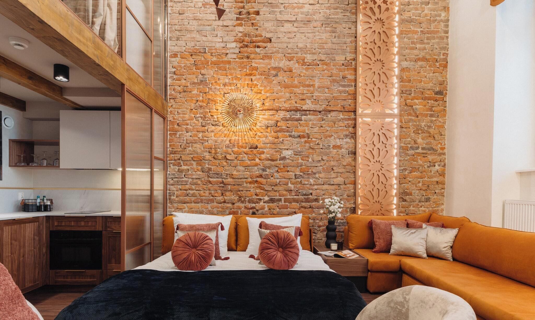 A modern room with a brick wall, a bed with orange bedding, a sofa, and a kitchen area with wooden cabinets.