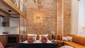 Een moderne kamer met een bakstenen muur, een bed met oranje beddengoed, een bank en een keuken met houten kastjes.