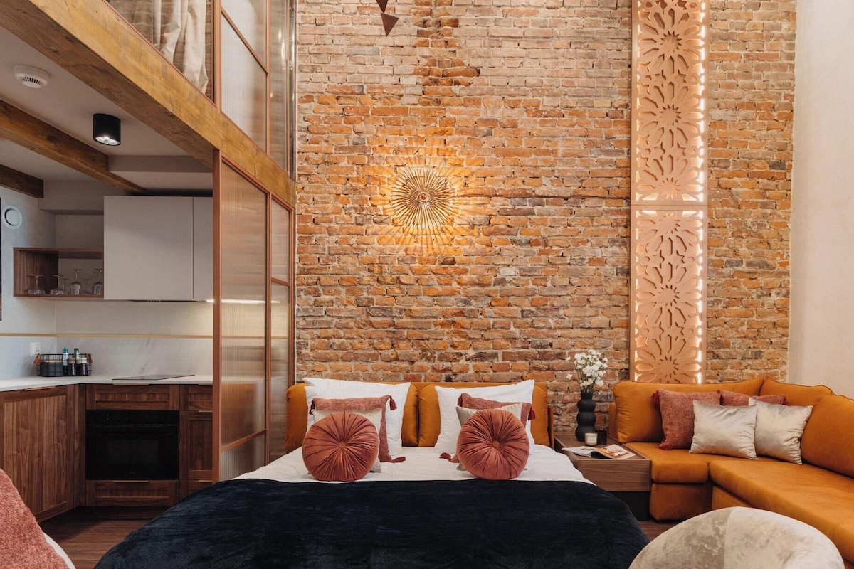 Una habitación moderna con pared de ladrillo, cama con ropa de cama naranja, sofá y una zona de cocina con armarios de madera.