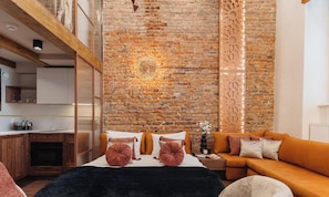 Una habitación moderna con pared de ladrillo, cama con ropa de cama naranja, sofá y una zona de cocina con armarios de madera.