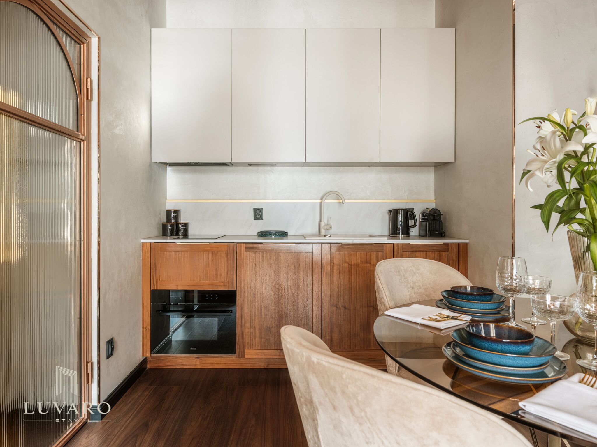 Una cocina moderna con armarios de madera, horno empotrado y comedor con mesa de vidrio para dos.