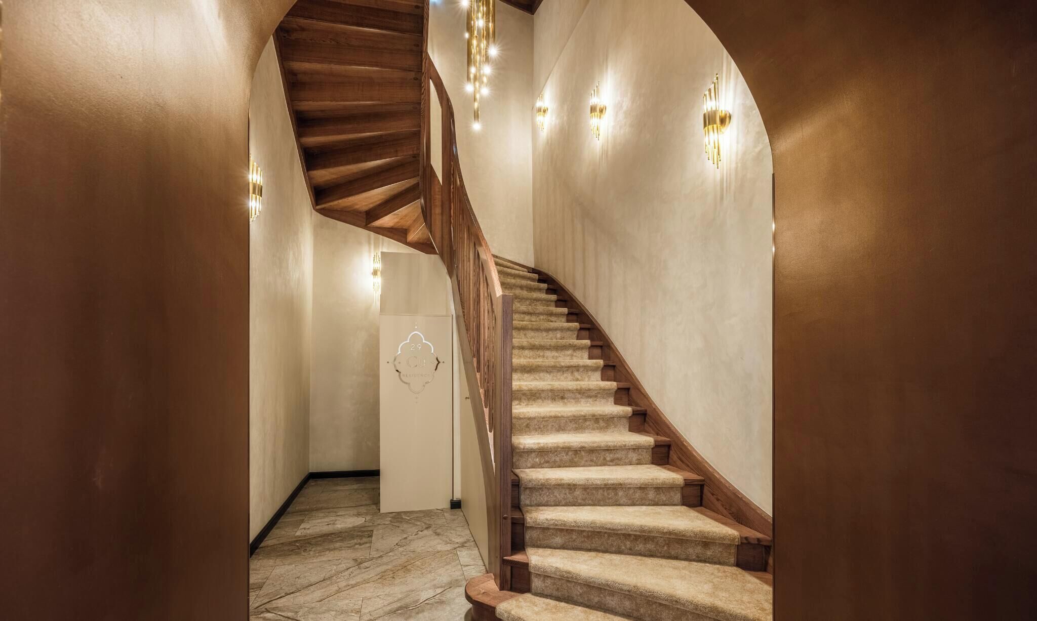 Una escalera de caracol con peldaños alfombrados y barandillas de madera, que conduce a una plataforma con una lámpara fijada a la pared.