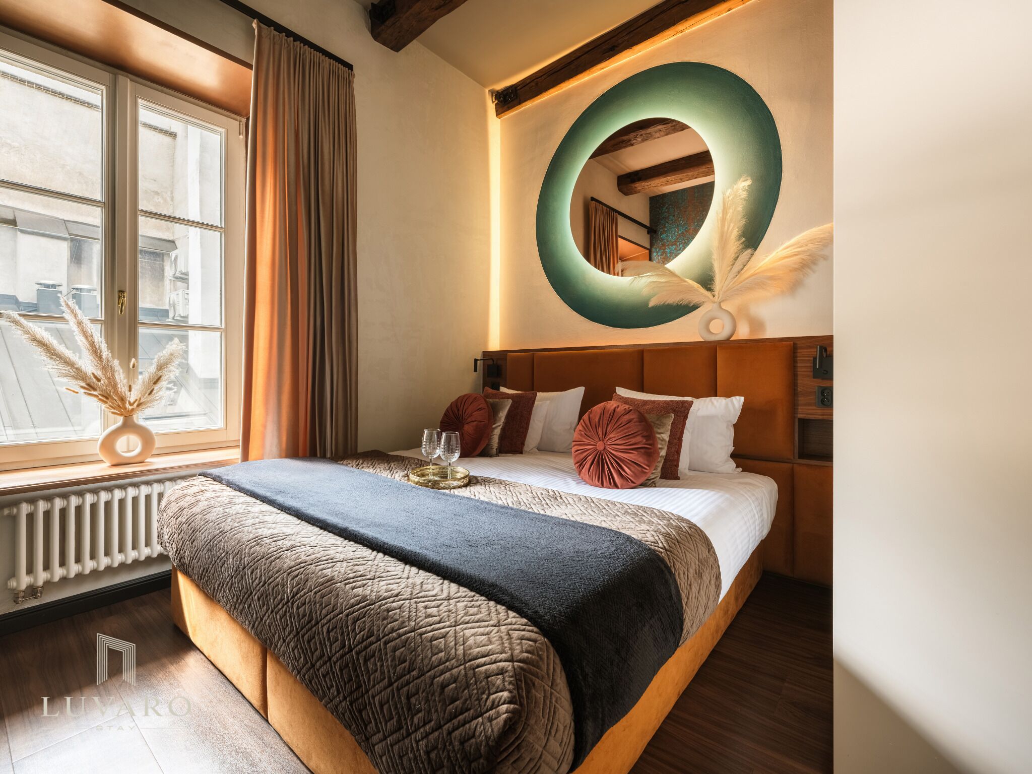 A hotel room with a large bed, a round mirror, and a view of the city through a window.