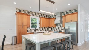 A modern kitchen with wooden cabinets, a central island with stools, a stainless steel refrigerator, and a hexagonal patterned backsplash. - Tango! Beach House for 26 Guests! Plus Golf Cart! (Santa Rosa Beach)